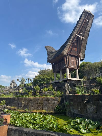 The image depicts a traditional Tongkonan house, a vernacular architectural style from Toraja, Indonesia, elevated on stilts and featuring a distinctive saddleback roof. This structure is situated in a landscaped garden environment. In the foreground, a pond filled with lily pads and lush green aquatic plants is visible. Behind the pond, a stone retaining wall creates tiered garden levels. Various plants, including ferns and grasses, grow along these terraces. A stone statue of a figure is positioned on one of the upper levels of the wall. In the background, other stone structures and landscaping elements are present, including ornate lamp posts. The sky above is blue with scattered white clouds. The scene suggests an outdoor setting designed to showcase architectural and botanical elements, likely within a park or botanical garden. This specific Tongkonan house replica is located at Pairi Daiza, a zoo and botanical garden in Cambron-Casteau, Belgium.