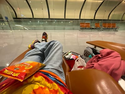 An individual is depicted from a first-person perspective, seated in a brown upholstered airport chair with their legs extended. The individual wears light blue denim jeans over a multi-colored tie-dye garment, and colorful athletic shoes. An unopened orange bag of "Gooral Crisp" potato chips rests on their lap. To the right, an adjacent brown chair holds a crumpled reddish-pink garment, a black and white checkered fabric item, a clear plastic water bottle, a white plastic bag, and other miscellaneous items within a red bag. The background shows a vast, brightly lit airport waiting area with a reflective floor and large glass panels. Multiple rows of modern brown-orange airport seating on metal frames are visible in the distance. The ceiling features a modern, curved architectural design with integrated lighting. This scene is located within Muscat International Airport (MCT) in Seeb, Seeb Province, Oman.