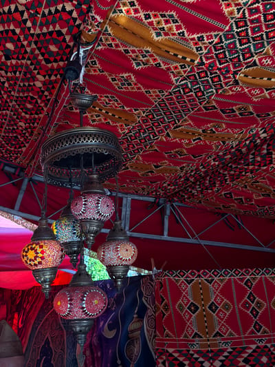 The image shows an interior scene, possibly within a tent or covered space, with an oriental or Middle Eastern aesthetic. Red and brown patterned fabrics are draped extensively, covering the ceiling and walls. A metal-framed structure is visible in the background. In the foreground, a chandelier-style light fixture with several individual lamps hangs. The lamps are decorated with colorful mosaics in various geometric patterns. The location is Burjassot, Spain.