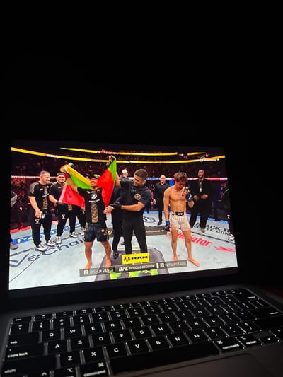 The image shows a laptop screen displaying a UFC (Ultimate Fighting Championship) event. On the screen, Joshua Van and Tatsuro Taira are identified.  A group of people is shown celebrating with a flag, possibly after a fight victory. A laptop keyboard is in the right foreground. The location is not mentioned in the image.