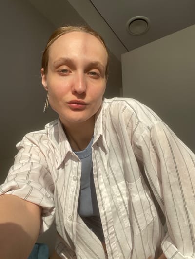 A woman is posing for a selfie indoors. She is wearing a striped white and grey button-down shirt over a blue top. The lighting is bright, casting shadows across the scene. In the background, there is a vent on the ceiling. Based on the available information, it is not possible to determine the exact location within Sepych, Russia.