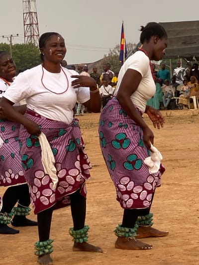 Two women in traditional African clothing dance. They wear white t-shirts, patterned purple skirts, black leggings, and spiky green ankle decorations. One holds a white cloth. Spectators sit in the background. A flag and a telecommunications tower are also visible.