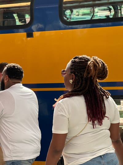 The image captures two individuals standing adjacent to a train in Lelystad, Netherlands. One individual, likely female, has braided hair with reddish-brown highlights pulled back into a bun, wears eyeglasses, and is adorned with gold earrings. She is wearing a white t-shirt and blue jeans. A gold chain is visible around her shoulder. The second individual, possibly male, is also wearing a white t-shirt and appears to have closely-cropped, dark hair. The background is dominated by a train with a blue and yellow exterior. A window and other design elements of the train are discernible. The scene appears to be outdoors in daylight.