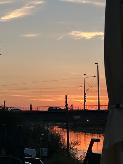 The media depicts a scene at dusk, likely in Mannheim, Germany, with the sky illuminated by the warm hues of sunset. The dominant elements are the sky, a bridge or overpass, and electrical infrastructure. The sky exhibits a gradient of soft oranges and yellows, with wispy clouds catching the last light of the day.
Below the sky, a dark structure resembling a bridge or elevated roadway spans across the frame, supported by columns. A metal railing runs along the edge of this structure. Visible on the right side of the bridge, a small, silhouetted figure stands, facing away from the camera, adding a sense of scale and human presence to the scene.
Electrical infrastructure, including poles and wires, crisscrosses the foreground and midground. Some poles are topped with lights, and a series of wires are strung between them. These structures suggest the presence of a transportation route, possibly a railway line, beneath or adjacent to the bridge.
In the lower right portion of the image, dark shapes of folded umbrellas and possibly seating suggest an outdoor seating area, perhaps a cafe or restaurant, implying a public space. The reflections on the water, which appears to be a river or canal, mirror the sunset colors, enhancing the atmospheric quality of the image. The overall mood is serene and contemplative, characteristic of a peaceful evening. No legible text is visible in the image.