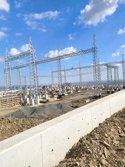 The image depicts a construction site for a high-voltage electrical substation, likely a part of the larger energy infrastructure development in Şereflikoçhisar, Turkey. Several lattice steel towers are erected on individual concrete foundations; wooden formwork is visible, suggesting ongoing concrete work. A concrete retaining wall runs along the foreground, with gravel and earth piled against it. In the background, there are earth-moving vehicles and further construction activity. The sky is blue with scattered cumulus clouds.