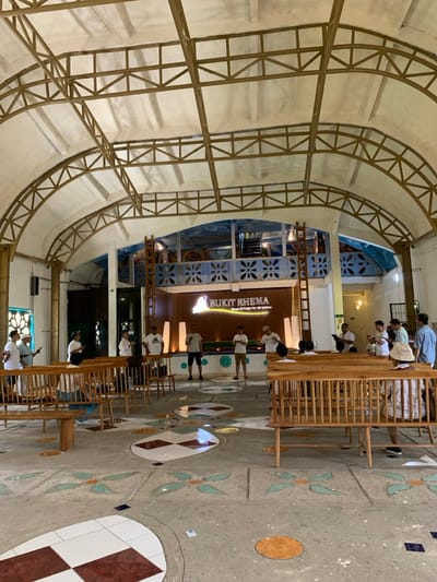 The image shows the interior of a large, open-air structure with a white concrete floor covered in mosaic tiles. The structure is supported by a network of brown metal beams forming an arched ceiling. The space is filled with wooden benches and several people are standing and sitting in various spots, some in discussion. There are a few people in the back of the room standing around a stage. The stage is elevated on a platform and decorated with blue painted walls with white floral patterns, and a banner that reads “BUKIT RHEMA”. There are a few potted plants and small lamps around the stage. The space is brightly lit, and the walls are white with a slight yellow tint. The image appears to have been taken in a large building or hall located at the coordinates -7.606, 110.181, which places it in Kecamatan Borobudur, Indonesia.