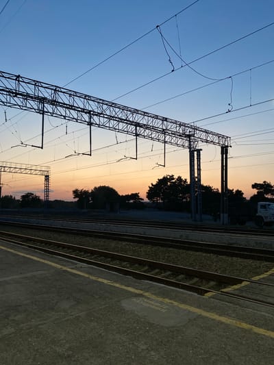 The image depicts a railway station at dusk in Samur, Russia. The sky is a gradient of deep blue transitioning to orange and pink near the horizon. Overhead, a complex network of overhead catenary wires and support structures for electric trains spans across the frame. In the middle ground, several sets of train tracks run parallel, bordered by a concrete platform in the foreground. Silhouetted trees and vegetation are visible in the background, with a large truck parked near the tracks on the right side. The platform edge is marked with a yellow safety line. The scene is devoid of any people or immediate action, conveying a sense of quiet anticipation.