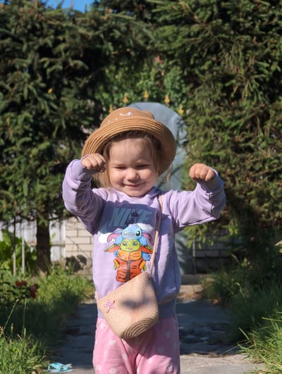 A young child stands outdoors in Perliavka, Ukraine, wearing a straw hat, a light purple hooded sweatshirt with a graphic print, and pink pants with white heart patterns. The child has their arms raised and fists clenched, appearing to be posing or making a playful gesture. A small, woven straw purse with a pink embellishment hangs from their shoulder. The background consists of dense green foliage, likely evergreen trees, under a bright blue sky. A stone pathway is visible in front of the child, with grass and some plants lining the sides. The lighting suggests it is daytime, with sunlight casting shadows.