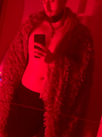 The image shows a person in a dimly lit room illuminated by red light. The person is wearing a fluffy, faux fur coat and appears to be holding a smartphone. They are also wearing a necklace with a cross pendant. The background is out of focus, but it appears to be a domestic setting. The image does not contain any information about the city of Gliwice, Poland.