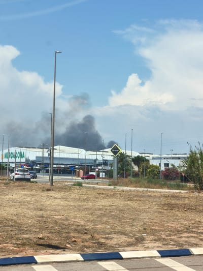 A photograph taken outdoors depicts a large commercial building with a white facade and a prominent green "MERCADONA" logo. Thick plumes of black smoke are rising from the roof of the building, indicating a fire. Several cars are parked in front of the building, and a red car is visible on a road nearby. A yellow diamond-shaped sign with the word "OLIVERAL" is also present. The scene is set against a blue sky with scattered clouds, and the surrounding area appears to be dry and grassy. The location is identified as Riba-roja de Túria, Spain, specifically near the Mercadona supermarket on Carrer dels Forners, as evidenced by the street sign.