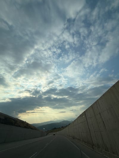 The image captures a view from a car driving on a highway through a mountainous region. The road is paved, with clear lane markings, and flanked by tall concrete retaining walls on both sides. The wall on the right is textured and angled upwards, while the wall on the left is partially obscured by vegetation and also angled. In the distance, rolling hills and mountains are visible, shrouded in a hazy atmosphere. The sky above is filled with a dramatic mix of light and dark clouds, with patches of blue sky peeking through, suggesting either dawn or dusk. The overall scene appears to be a scenic drive, possibly in a rural or semi-urban area outside of a major city, consistent with the outskirts of Muğanlı, Azerbaijan, which is known for its mountainous terrain and highways. There are a few vehicles visible on the road, indicating active traffic. No people are directly visible within the frame.