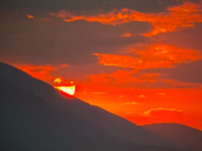 The media file captures a dramatic sunset over a mountainous landscape in Kovachevets, Bulgaria. The sun, a brilliant orb of intense orange and yellow, is partially obscured by the silhouette of a mountain range, casting a fiery glow across the sky. Wispy clouds, illuminated by the setting sun, drift across the horizon, adding depth and texture to the scene. The foreground is dominated by the dark, imposing shapes of the mountains, creating a stark contrast with the vibrant sky. The overall impression is one of natural beauty and tranquility, with the vibrant colors evoking a sense of awe and wonder. The time of day is clearly sunset, indicated by the low position of the sun and the warm, intense hues of the sky. No people or objects are visible in the frame, and there is no visible text. The location cues suggest a rural or mountainous setting.