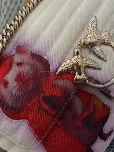 The image displays a close-up of a white quilted bag with a decorative chain and bird embellishments. Two golden bird charms are prominently featured on the bag's surface. One bird is in the foreground, its wings spread wide, while another is slightly behind it and to the left. A circular silver clasp attaches the birds to the bag. The bag itself has a textured, quilted pattern with subtle white stitching.
Overlaid on the white fabric are printed graphics in shades of red, purple, and black. These graphics appear to be abstract or artistic, with one section resembling the face of an animal, possibly a cat or a fox, due to the visible eyes and muzzle-like features. The prints have a slightly translucent quality, allowing the underlying quilting to show through.
To the left of the frame, a portion of a thick, silver metal chain is visible, suggesting it might be a strap or handle for the bag. The background is out of focus but appears to be a striped textile, possibly bedding or upholstery. The lighting suggests an indoor setting, likely during the day, with no strong shadows indicating a specific time. The overall aesthetic is one of fashion and accessory detail, with a focus on the intricate craftsmanship of the bag and its decorative elements. There is no visible text in the image.