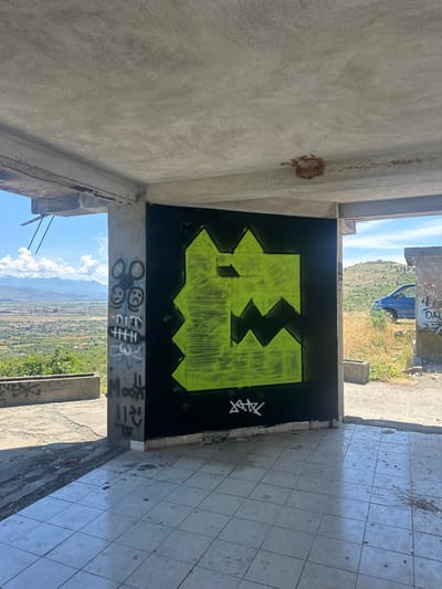 This image depicts the interior of a partially constructed or abandoned building in Gornji Kokoti, Montenegro. The foreground is dominated by a tiled floor with some debris. On the back wall, a large, jagged graffiti tag in bright lime green spray paint is prominently displayed against a black background. To the left of the graffiti, a concrete pillar is covered in various smaller tags and drawings, including smiley faces and what appears to be the word "Moon".

The building appears to be open to the elements, with views of a rural landscape in the background. Rolling hills covered in vegetation, a town or village, and mountains under a bright blue sky with scattered clouds are visible. A blue car is parked outside the structure to the right. The overall scene suggests a state of disrepair and neglect, common in abandoned or unfinished structures.