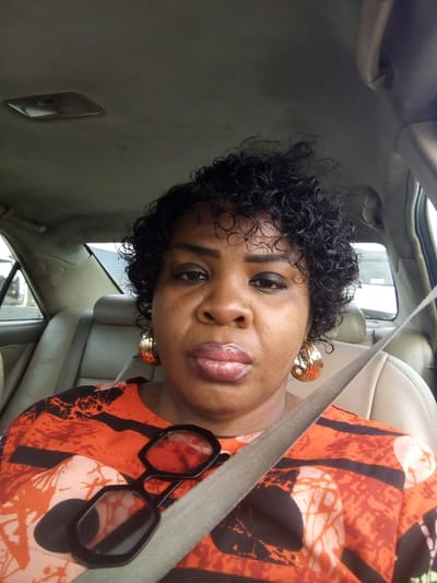 A woman sits in a car, wearing a patterned orange and black top, secured by a seatbelt. Black-rimmed glasses rest on her chest, and she sports large, gold hoop earrings. Her hair is short, dark, and curly. The car interior is light-colored, with the back seats visible. The woman gazes directly at the camera with a neutral expression. A window is seen behind her.
