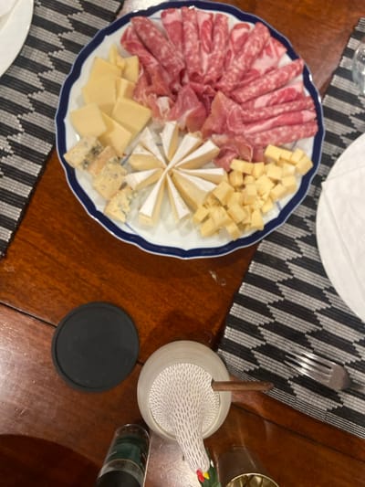 A white plate with a blue rim containing a cheese and meat platter is on a wooden table, situated in Rye, New York. The plate is centered on a black and white patterned placemat. The platter includes sliced salami and prosciutto, a wheel of brie, cubed cheddar cheese, and two types of blue cheese. A black round coaster is to the left of the plate. To the right of the plate is a white placemat with a silver fork on top. A ceramic chicken figurine is below the placemat, and a glass jar containing a spice blend is in the bottom left corner.