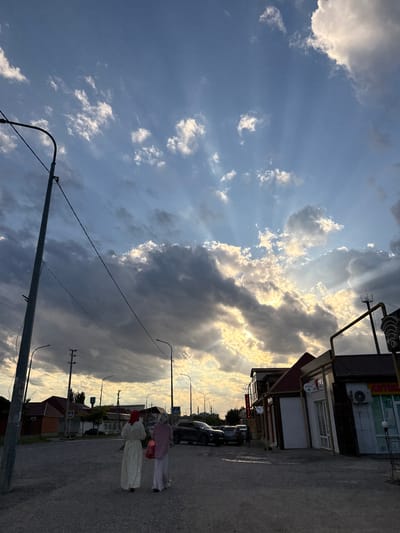 Two women are walking on a paved street in Kurchaloy, Russia, under a dramatic sky. The sky is filled with a mix of dark, heavy clouds and bright, sunlit patches, with rays of sunlight streaming through. This suggests it is either sunrise or sunset. The buildings lining the street appear to be commercial establishments, with one clearly marked as "АПТЕКА" (Apteka, meaning pharmacy) and another with Cyrillic text that is partially obscured. There are cars parked along the side of the road. The overall atmosphere is serene, with the interplay of light and shadow creating a visually striking scene.