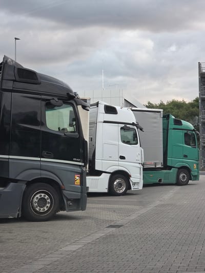 Three semi-trucks are parked side-by-side in a paved lot. The truck on the left is black and gray, with a white stripe along its side. The middle truck is white. The truck on the right is green. All three trucks have their trailers attached. The lot is made of gray brick pavers. In the background, there is a building with a modern design, trees, and a metal structure with stairs. The sky is overcast with clouds. A streetlamp is visible in the upper left corner of the image. A sign that says "ATTENTION ANGLES MORTS" is visible on the lower part of the black and gray truck. The location is identified as Zeppelinheim, Germany.