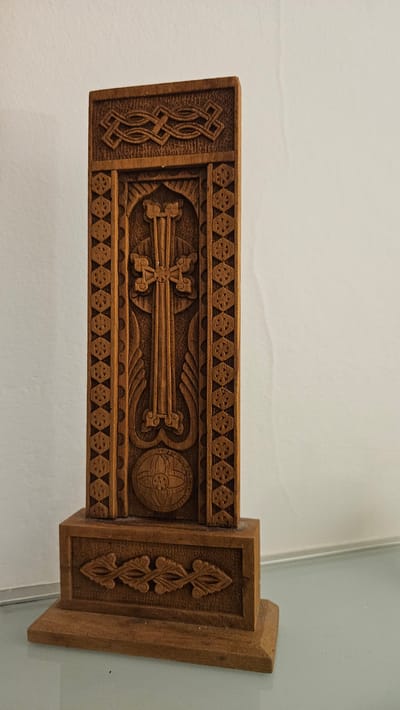 A wooden, carved object is displayed on a light-colored surface, against a white background. The object is a vertical piece with intricate carvings. At the top, there is a horizontal band with a knot-like pattern. Below this, the main body of the object features a stylized cross as the central element, flanked by vertical decorative bands. Beneath the cross is a circular carving with geometric patterns, possibly representing a globe or a stylized sun. The object is mounted on a square base, which is adorned with a horizontal carved band featuring a floral or leaf-like motif. The carving is detailed and appears to be made of a medium-toned wood. The lighting casts subtle shadows, highlighting the texture and depth of the carvings. The scene suggests an indoor setting, with the object likely being a decorative item or a religious artifact.