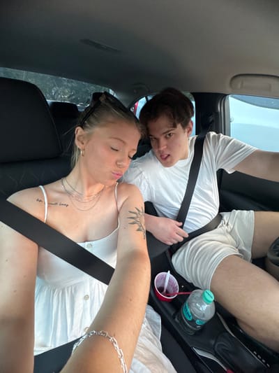 A young couple is in a car. The woman is in the foreground wearing a white dress and a seatbelt. She has light hair and tattoos on her arm and chest. The man is in the background, also wearing a white shirt and seatbelt. He has dark hair and is sticking his tongue out. A red cup with a straw, a water bottle, and some white fabric are visible in the background. The photo is taken from above, looking down into the car.