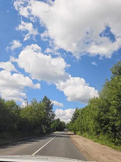 The image captures a view from inside a vehicle, looking forward along a two-lane asphalt road. The road has a white line dividing the lanes. Lush green trees line both sides of the road, with a sandy, uneven shoulder on the right. In the distance, the road curves slightly to the left, and a few trucks are visible. The sky above is a bright blue, adorned with fluffy white clouds. The overall scene suggests a clear, sunny day in a rural or semi-rural setting, possibly on the outskirts of Торбеево, Russia, given the context.