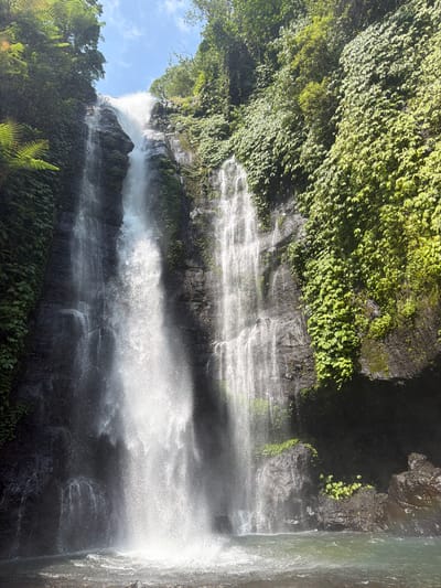 A tall, multi-tiered waterfall cascades down a dark, moss-covered rock face in a lush, verdant environment. The water is white and frothy, with a fine mist rising from the impact at the bottom. The surrounding cliffs are covered in dense green foliage, including ferns and vines, with trees visible at the top. The water collects in a clear, tranquil pool at the base of the falls. The sky above is a bright blue with scattered white clouds. This is the Banyumala Waterfall in Sawan, Indonesia, a natural wonder known for its beauty and serene atmosphere.
