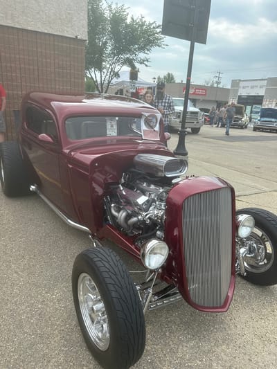 This image captures a street scene in the City of Leduc, Canada, likely during an outdoor event or car show. The primary subject is a prominently featured, customized maroon hot rod, showcasing its exposed engine, chrome accents, and wide tires.
Several people are visible in the background, suggesting a gathering or public event. Some individuals are casually walking, while others appear to be browsing or interacting near tents and vehicles. The setting includes paved ground, buildings with visible signage, and trees, indicating an urban or town environment. The sky is overcast, suggesting a cloudy day, and the lighting implies it's daytime, likely late morning or afternoon.
Notable details include the "Miller Brewing Company" text visible on a building, a "Highway 30" sign, and a "30" speed limit sign, providing specific location context. The hot rod itself is a significant focal point, with its powerful engine and polished exterior. The overall mood seems relaxed and social, typical of a community event.