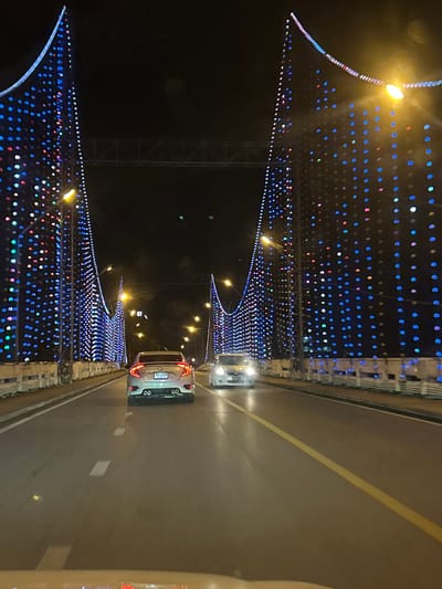 The image captures a nighttime scene on a road decorated with numerous colorful lights. The perspective is from within a vehicle, looking forward and slightly to the left. The road itself is asphalt, with a prominent yellow lane divider. On the left side of the road, a structure adorned with a dense pattern of blue, white, and some red lights is visible. These lights form an elaborate display that appears to be part of a bridge or a similar architectural feature. The lights are arranged in rows and create a festive, almost magical ambiance.
Further down the road, another vehicle, a silver car, is driving ahead. Its taillights are illuminated, and its license plate is visible, though the text is too blurry to read. The car appears to be a sedan or a hatchback.
The overall setting is a road at night, likely in an urban area, given the extensive light decorations. The lights suggest a celebratory occasion or a permanent decorative installation in Surat Thani City Municipality, Thailand. The darkness of the night is contrasted by the vibrant artificial lights, creating a visually striking scene. There is a sense of movement as the vehicles are on a road, implying travel or transit. The weather appears clear, as there are no indications of rain or fog. The mood conveyed by the lights is cheerful and lively.
