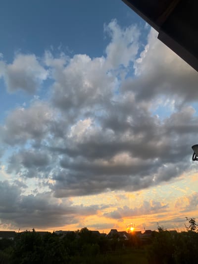 The image depicts a twilight sky with dramatic cloud formations over a rural landscape. The sun is setting on the horizon, casting a warm orange and yellow glow across the sky and illuminating the undersides of the clouds with a bright, golden light. The clouds themselves are a mix of dark gray and white, with some appearing heavy and textured while others are wispy and thin.
In the foreground, a silhouetted line of trees and rooftops suggests a small settlement or village. The architecture of the buildings appears simple, consistent with a rural setting. The vegetation is dense and dark, with the green hues muted by the fading light. A dark, angled structure, possibly part of a roof or overhang, occupies the upper right portion of the image, framing the sky. The overall impression is one of a serene, yet dynamic, natural scene at the end of the day. There are no people or discernible actions taking place in the image; it is a still landscape view.