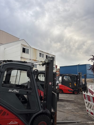 An outdoor image depicts a lot with multiple forklifts and stacked shipping containers under a cloudy sky. In the foreground, a dark gray and red Linde forklift is partially visible, with its "Linde" logo clearly displayed on the lower red portion. To its right, a red and black forklift with "Jungheinrich" branding is parked. Further back, a row of white shipping containers, some stacked two high, are visible, featuring numerous windows. A large blue building with a corrugated metal facade stands to the right of the containers. The ground is paved with asphalt and shows white markings. There are no people visible in the image. The scene appears to be an industrial or storage area.