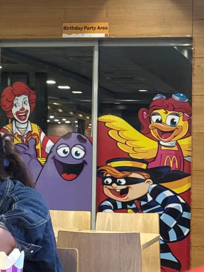 The image depicts a colorful mural on a glass partition, likely in a fast-food establishment like McDonald's, given the presence of Ronald McDonald and other cartoon characters. The mural features several cartoon characters, including a character resembling Birdie the Early Bird from McDonald's, with yellow feathers and a pink outfit, and another character in a black and blue checkered outfit, possibly Grimace or a similar mascot. Below this, there's a depiction of Ronald McDonald himself, with his signature red hair and white face paint, alongside a purple, blob-like character with large eyes. The mural has a festive and playful appearance, suggesting a child-friendly environment.

A sign on the left side of the partition reads "Birthday Party Area" in English, reinforcing the notion that this is a designated space for celebrations. The lighting suggests it's daytime, with bright interior lights reflecting on the glass. The background seen through the glass appears to be the interior of a restaurant, with some blurry shapes and lights visible. A person wearing a denim jacket is partially visible in the foreground, obscuring some of the lower part of the mural. The overall scene conveys a cheerful and energetic atmosphere, characteristic of a fast-food restaurant's play or party area. The location context is Manila, Philippines.