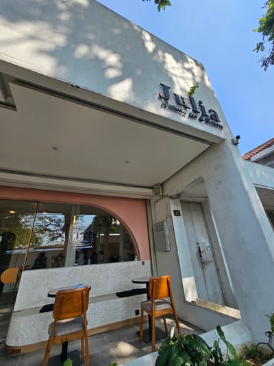 A low-angle shot captures the exterior of a building with the name "Julia" prominently displayed in black lettering on the facade. Below "Julia" is smaller text reading "Artisan tea & Bakery". The building appears to be a commercial establishment, possibly a cafe or bakery, given the signage. The facade is a light-colored concrete, with a portion of a lighter-colored awning or overhang visible at the top. There is a curved section of the building to the left, with a large window that reflects trees and the sky. Inside the window, some tables and chairs are visible, along with a sign that says "Julia's".
To the right of the entrance, there is a closed white door. Next to the door, there's a gray electrical box with three switches. Above the door, a security camera is mounted. A sign with the number "28" is affixed to the wall near the door.
In front of the building, there is outdoor seating with two small round tables and two chairs. The chairs are made of wood with gray upholstered seats. A built-in bench made of speckled concrete runs along the front of the building, also providing seating. Lush green plants are visible in the foreground, partially obscuring the base of the building and the outdoor seating area.
The sky is clear and blue, with dappled sunlight casting shadows from unseen trees onto the building's facade. The image is taken on a sunny day, suggesting warm weather. The architectural style is modern and minimalist, with clean lines and a neutral color palette. The location is identified as Banyuurip, Indonesia.
