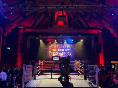 A boxing ring occupies the center of a room, with a camera focused on it. A screen displaying "FIGHT NIGHT 2025" is visible behind the ring. People surround the ring, some standing and others seated. Lighting rigs hang overhead, illuminating the space with a mix of spotlights and colored lights, primarily red and blue. The ornate architecture of the room adds a sense of grandeur to the event.