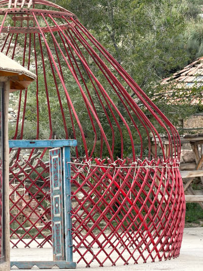 The image captures a partially constructed yurt in the village of Kyzyl-Tuu, Kyrgyzstan. The yurt's wooden frame, painted red, is visible, with latticed lattice walls tied together with white rope. Several red wooden poles radiate upwards from the base, forming the ribs of the yurt. A section of a blue-painted wooden door with decorative patterns is propped open, revealing more of the lattice structure behind it. In the background, a tiled roof of a building is visible, along with green foliage and trees. The ground is a light-colored gravel or dirt surface.