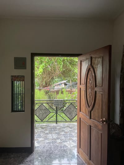 An interior view looks out through an open wooden door to a lush, green outdoor area. To the left of the door is a narrow window with a metal grate, topped by a colorful decorative fabric. Above the window, mounted on the white wall, is a gray electrical box with an orange switch. The floor inside is tiled with large, dark, glossy tiles that reflect the light.

The open doorway reveals a small, tiled patio with irregularly shaped, multi-colored tiles. A metal railing with diamond-shaped patterns runs along the edge of the patio. Beyond the railing, a dense canopy of green trees and foliage dominates the scene, with hints of a small structure, possibly a shed or shack, visible in the background. The overall atmosphere is one of a simple dwelling opening onto a verdant, tropical environment in Bacong, Philippines.