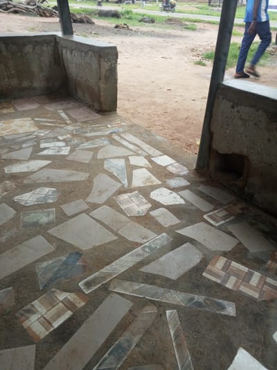 The image depicts an outdoor scene with a focus on a floor paved with irregularly shaped stones and some decorative tiles. The paving appears to be an unfinished or rustic construction, with sand filling the gaps between the stones and tiles. In the background, there is a dirt path and some vegetation, suggesting a rural or semi-urban environment. A person, partially visible, is walking away from the camera on the dirt path. The scene is located in Gindin Dutse, Nigeria.
