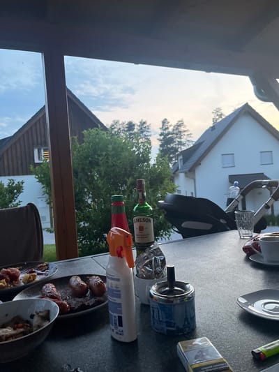 This image depicts an outdoor gathering at dusk, likely in a residential area given the presence of houses in the background. The setting is a patio or balcony table, cluttered with items suggestive of a casual meal or drinks.
Prominently featured on the table are several bottles, including a green bottle of Jameson Irish Whiskey, and what appears to be a red bottle. A spray bottle with an orange nozzle is also visible, along with an ashtray containing cigarette butts. The scene includes a small, clear glass, and a cigarette pack. Plates with food, possibly cooked meats or sausages, are arranged on the table, indicating a meal is being consumed or has just finished.
In the background, a white house with dark roofs and windows is visible, partially obscured by green foliage. A baby stroller is also present, suggesting the presence of a family. The sky is visible, indicating it is late afternoon or early evening, with a warm, diffused light suggesting either sunrise or sunset. The overall mood is relaxed and informal, hinting at a social gathering. The image is oriented sideways, with the horizon appearing vertical.
Visible text includes "JAMESON TRIPLE DISTILLED IRISH WHISKEY" on the Jameson bottle. The cigarette pack has text that is partially obscured but appears to be a brand name. The spray bottle has a label with text and graphics that are too small to decipher clearly. The location context provided is Milná, Czechia.