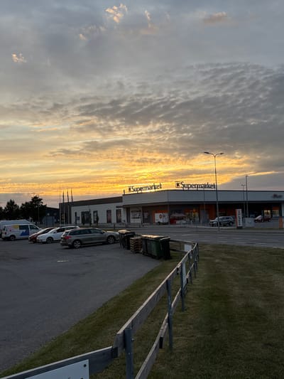 An exterior, low-angle, medium shot captures a supermarket in Pori, Finland, under a dramatic sky at sunset. The sun, a brilliant orange and yellow, illuminates a vast expanse of clouds, casting a warm glow across the scene. The K-Supermarket building, with its prominent signage, stands in the background. Several cars are parked in the lot, including a white van, a red car, a white sedan, and a silver station wagon. In the foreground, a rustic wooden fence borders a grassy area. The overall impression is one of a peaceful, late evening in a Finnish city.