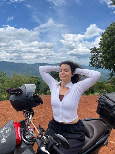 A young woman with curly dark hair and closed eyes sits on a motorcycle, resting her hands behind her head. She wears a white zip-up jacket over a brown top and black pants. She has black earbuds in her ears. A black motorcycle helmet with brown fur ear covers and a black bow is on the handlebars. A red water bottle with "SCITEC NUTRITION" is attached to the motorcycle. The motorcycle is parked on a reddish-brown dirt surface. In the background, lush green hills and mountains are visible under a partly cloudy blue sky. The scene is likely in a rural mountainous area, given the landscape.