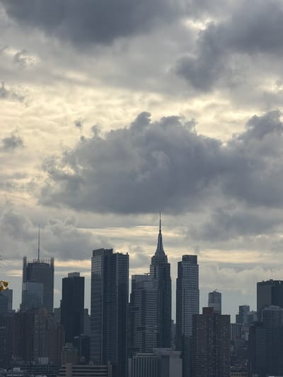 A static, panoramic view captures the Manhattan skyline under a dramatic, heavily overcast sky, observed from an elevated vantage point in Weehawken, United States. The upper two-thirds of the frame are dominated by a dense formation of dark grey and lighter cumulus clouds, with brighter, diffused sunlight visible through a horizontal band of thinner cloud cover in the mid-sky. The lower third of the image features a concentrated cluster of high-rise buildings. The Chrysler Building, identifiable by its distinct conical, spired apex, is prominently positioned slightly right of the frame's vertical center. To its left, a tall, rectangular skyscraper exhibiting an antenna or spire and visible scaffolding is present. Numerous additional modern glass and steel towers, alongside older, darker-toned buildings, collectively form the extensive cityscape. No individuals, vehicles, or specific ground-level activities are discernible within the frame.