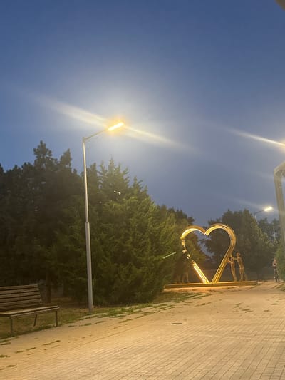 In the city of Ilyinsky, Russia, a twilight scene unfolds in a park setting, bathed in the soft glow of streetlights. Dominating the midground is a large, illuminated heart-shaped sculpture, fashioned from what appears to be gold-toned metal. Within the heart, two stylized human figures are depicted, seemingly interacting. To the left of the sculpture, a dense cluster of evergreen trees forms a lush backdrop. In the foreground, a paved walkway, composed of light-colored rectangular bricks, stretches into the distance. A weathered wooden bench, with a slatted seat and backrest, sits to the left of the walkway. A tall streetlight stands to the left of the bench, its bright light casting a glare across the image. Further to the right, another streetlight is visible, and in the far distance on the right side of the walkway, a person on a bicycle or scooter is present. The sky above is a deep blue, transitioning to a lighter hue near the horizon, with some faint cloud formations or atmospheric effects visible.
