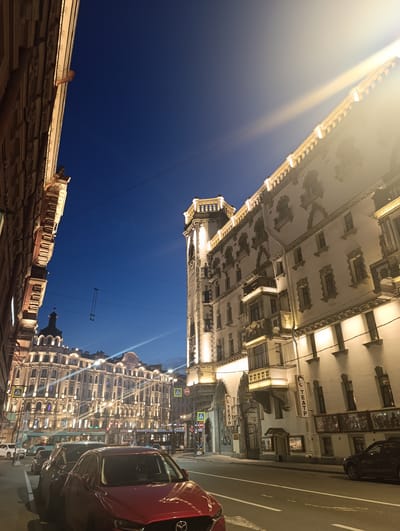 The image captures a street scene in Saint Petersburg, Russia, at dusk. The architecture suggests a historic district, with ornate buildings lining a wide street. The buildings are illuminated, highlighting their detailed facades and numerous windows. A prominent tower-like structure is visible on the right, with decorative elements and warm lighting. Several cars are parked along the street and in motion, including a red car in the foreground and a black car on the right. Pedestrian crossing signs are visible, indicating a public area. The sky is a deep blue, transitioning from twilight. The overall atmosphere is that of a well-lit urban environment at the end of the day.