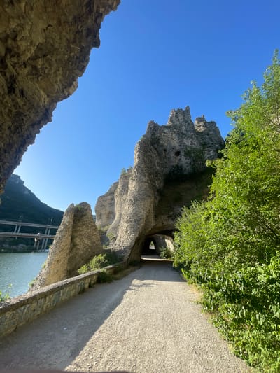 The image depicts a scenic view of a rocky formation and a pathway near a body of water. The rocky formations are rough and jagged, with some appearing to be carved or shaped. Lush green foliage spills over the tops of these rocks, suggesting a natural or semi-natural environment.
A paved pathway runs alongside the rocky formations, curving gently. This pathway is bordered by a low stone wall. In the distance, a tunnel-like opening is visible within the rock, leading to darkness. Beyond the pathway and rocks, a wide body of water, possibly a river or lake, is visible. Across the water, a lush green mountainside rises, with a bridge structure supported by multiple pillars spanning the water and extending towards the mountain. The sky is a clear, vibrant blue, indicating a sunny day. The angle of the sun suggests it is either morning or late afternoon.
The scene evokes a sense of exploration and natural beauty. The combination of rugged rocks, vibrant greenery, and the serene water creates a peaceful yet adventurous atmosphere. There is no visible text in the image. The location is identified as Asparuhovo, Bulgaria.