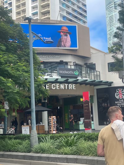 An outdoor, eye-level, medium shot captures a street scene in Surfers Paradise, Australia. In the background, a large digital billboard displays a close-up of a bird wearing a straw hat and sunglasses, with text that partially reads "ch, t fraud." Below the billboard, the facade of a building features prominent signage for "SPORTING GLOBE BAR & GRILL" and "WOOLWORTHS." The lower portion of the image shows lush green foliage and a street-level view of people walking and sitting. A grey lamppost with a decorative base stands in the foreground, slightly off-center. The sky is partly cloudy.