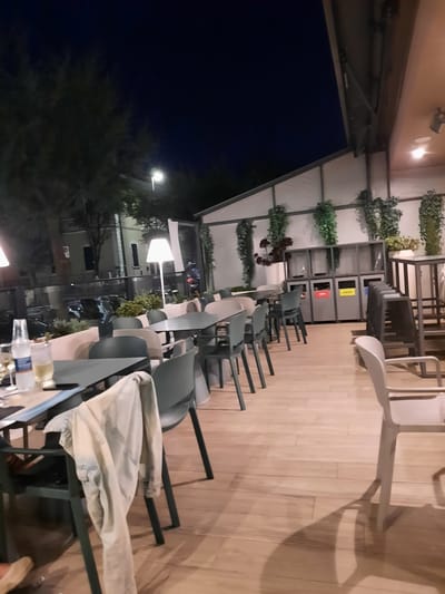This is an exterior shot of an outdoor seating area at night, likely a restaurant or cafe, in Pesaro, Italy. The area is furnished with multiple tables and chairs. In the foreground, a table is set with a glass of white wine and a bottle of water. A light-colored jacket or garment is draped over the back of a chair. In the background, there are trees and buildings visible against a dark night sky. A street lamp illuminates the scene. Towards the right side of the image, there are recycling bins with labels in Italian. The floor is made of wooden planks. The overall atmosphere suggests a quiet evening, with the establishment closed or preparing to close.