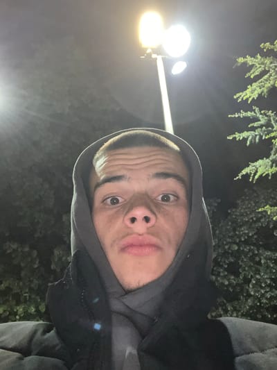 A selfie of a young man with light skin and short hair, wearing a hooded sweatshirt, is taken outdoors at night. A street light is visible in the background. Vegetation is also visible in the background. The location is not evident from the image.