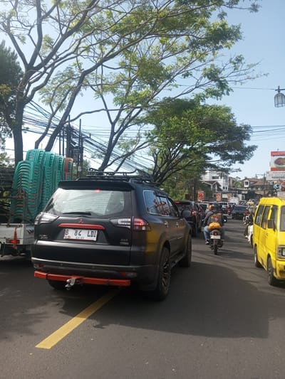 The image shows a street in what appears to be a busy urban area with a high volume of traffic. The sky is clear and blue. Several trees with green leaves line the road. Various vehicles are visible, including a dark-colored SUV with a roof rack and a red bumper. To the left of the SUV, there is a truck loaded with stacked green plastic chairs. A yellow minivan is visible to the right of the SUV. Several motorcycles are in the lane with other vehicles, and several buildings and advertisements can be seen in the background. The license plate on the black SUV reads "B 84 LDI 01-31 4X4". The location is not Khe Luang, Laos. Signs in the background appear to be in Indonesian.