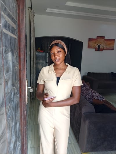 A young woman stands at a doorway in what appears to be a residential interior in Basawa, Nigeria. She is wearing a tan jumpsuit with a black top underneath and a tan headband. She holds a pink smartphone in her left hand. The doorway opens into a living room with a dark gray sofa and a person lying on it wearing a patterned dress. On the wall behind the sofa hangs an abstract painting of a tree. The door itself has a textured gray panel design with a brown frame and a silver handle.