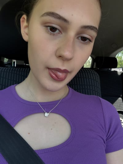 A close-up, tilted selfie of a young woman in a car, presumably in Levski, Bulgaria. She has fair skin, brown eyes, and arched eyebrows. She is wearing a purple ribbed top with a cutout near the collarbone and a silver necklace with a heart-shaped pendant. A black seatbelt is visible across her chest. The car seat is upholstered in a dark gray patterned fabric. Through the car window, a glimpse of greenery and trees can be seen. Her lips are glossy, and her tongue is slightly visible.