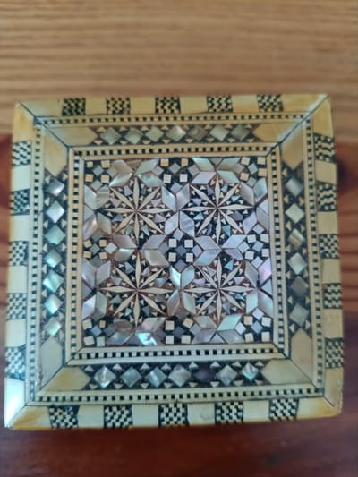 A close-up, overhead shot shows the intricate inlay work on the lid of a wooden box, placed on a wooden surface. The box lid features a central square pattern composed of mother-of-pearl pieces in geometric shapes, primarily diamonds and eight-pointed stars, interspersed with dark tiles and small squares. Surrounding this central design is a border of lighter wood with inlaid geometric patterns, including a band of small, alternating black and white squares, followed by a diamond pattern within a light frame, and further bands of geometric inlay. The outer edges of the box lid are also adorned with the black and white checkered pattern. The wooden surface beneath the box displays a natural grain pattern. The image does not contain any people or specific city landmarks.